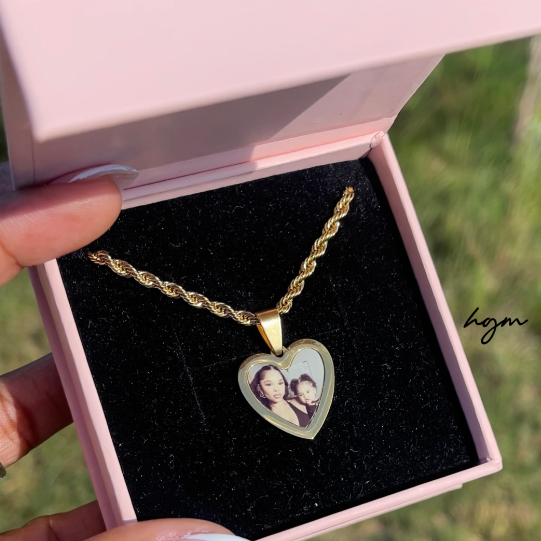 Gold heart-shaped photo necklace with custom picture pendant on a rope chain, displayed in a pink jewelry box held outdoors in natural light.